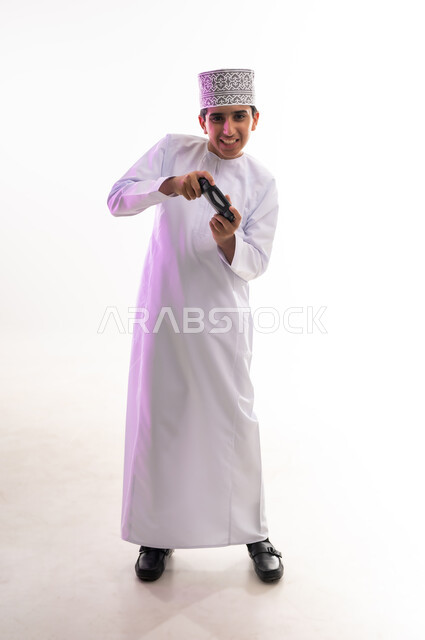 قضاء أوقات ممتعة في ألعاب الفيديو الترفيهيه، بورتريه لولد عربي خليجي عماني يرتدي الدشداشة والكمة يحمل بيده عصا التحكم، إيماءات الاستمتاع والاندماج، استخدام الأجهزة التكنولوجية والتقنيات الحديثة، خلفية بيضاء