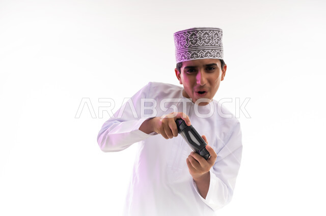قضاء أوقات ممتعة في ألعاب الفيديو الترفيهيه، بورتريه لولد عربي خليجي عماني يرتدي الدشداشة والكمة يحمل بيده عصا التحكم، إيماءات الاستمتاع والاندماج، استخدام الأجهزة التكنولوجية والتقنيات الحديثة، خلفية بيضاء