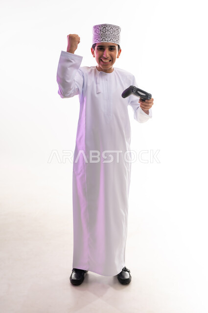قضاء أوقات ممتعة في ألعاب الفيديو الترفيهيه، بورتريه لولد عربي خليجي عماني يرتدي الدشداشة والكمة يحمل بيده عصا التحكم، إيماءات الاستمتاع والاندماج، استخدام الأجهزة التكنولوجية والتقنيات الحديثة، خلفية بيضاء