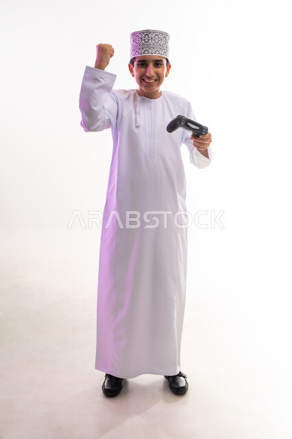 قضاء أوقات ممتعة في ألعاب الفيديو الترفيهيه، بورتريه لولد عربي خليجي عماني يرتدي الدشداشة والكمة يحمل بيده عصا التحكم، إيماءات الاستمتاع والاندماج، استخدام الأجهزة التكنولوجية والتقنيات الحديثة، خلفية بيضاء