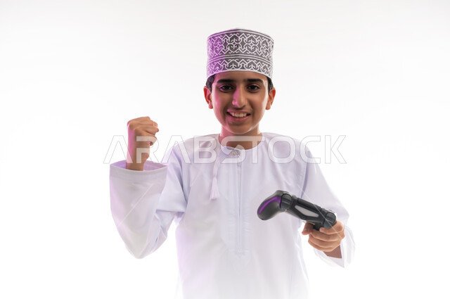 قضاء أوقات ممتعة في ألعاب الفيديو الترفيهيه، بورتريه لولد عربي خليجي عماني يرتدي الدشداشة والكمة يحمل بيده عصا التحكم، إيماءات الاستمتاع والاندماج، استخدام الأجهزة التكنولوجية والتقنيات الحديثة، خلفية بيضاء