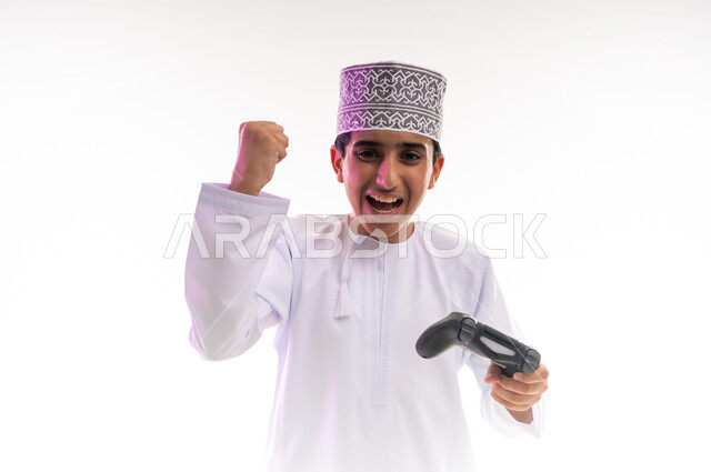 قضاء أوقات ممتعة في ألعاب الفيديو الترفيهيه، بورتريه لولد عربي خليجي عماني يرتدي الدشداشة والكمة يحمل بيده عصا التحكم، إيماءات الاستمتاع والاندماج، استخدام الأجهزة التكنولوجية والتقنيات الحديثة، خلفية بيضاء