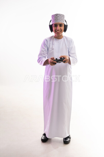 Artistic singing talents, interaction with musical tunes, the concept of listening and enjoying, listening to songs using headphones, spending fun times playing video games, a portrait of an Arab Gulf Omani boy wearing a dishdasha and a turban, wearing headphones and holding a joystick in his hand, white background