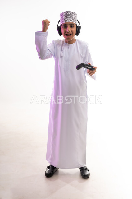 مواهب غنائية فنية، التفاعل مع الأنغام الموسيقية، مفهوم الاصغاء والاستمتاع، الاستماع للأغاني باسخدام الهيدفون، قضاء أوقات ممتعة في ألعاب الفيديو الترفيهيه، بورتريه لولد عربي خليجي عماني يرتدي الدشداشه والكمه يضع سماعات الرأس يمسك بيده عصا التحكم,  خلفيه بيضاء