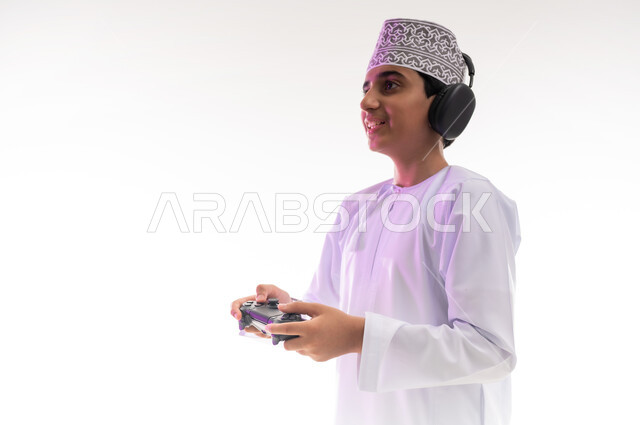 Artistic singing talents, interaction with musical tunes, the concept of listening and enjoying, listening to songs using headphones, spending fun times playing video games, a portrait of an Arab Gulf Omani boy wearing a dishdasha and a turban, wearing headphones and holding a joystick in his hand, white background