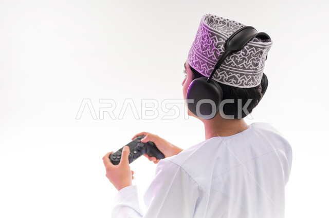مواهب غنائية فنية، التفاعل مع الأنغام الموسيقية، مفهوم الاصغاء والاستمتاع، الاستماع للأغاني باسخدام الهيدفون، قضاء أوقات ممتعة في ألعاب الفيديو الترفيهيه، بورتريه لولد عربي خليجي عماني يرتدي الدشداشه والكمه يضع سماعات الرأس يمسك بيده عصا التحكم,  خلفيه بيضاء