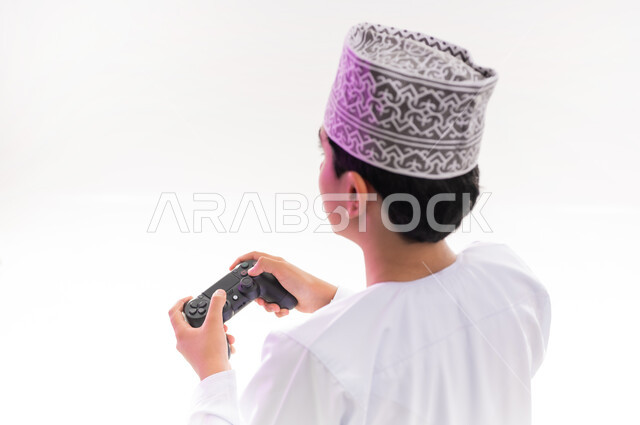 قضاء أوقات ممتعة في ألعاب الفيديو الترفيهيه، بورتريه لولد عربي خليجي عماني يرتدي الدشداشة والكمة يحمل بيده عصا التحكم، إيماءات الاستمتاع والاندماج، استخدام الأجهزة التكنولوجية والتقنيات الحديثة، خلفية بيضاء