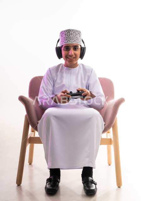مواهب غنائية فنية، التفاعل مع الأنغام الموسيقية، مفهوم الاصغاء والاستمتاع، الاستماع للأغاني باسخدام الهيدفون، قضاء أوقات ممتعة في ألعاب الفيديو الترفيهيه، بورتريه لولد عربي خليجي عماني يرتدي الدشداشه والكمه يضع سماعات الرأس يمسك بيده عصا التحكم,  خلفيه بيضاء