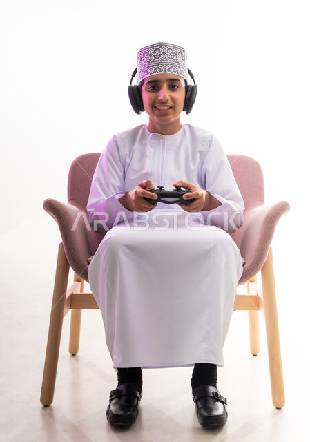 Artistic singing talents, interaction with musical tunes, the concept of listening and enjoying, listening to songs using headphones, spending fun times playing video games, a portrait of an Arab Gulf Omani boy wearing a dishdasha and a turban, wearing headphones and holding a joystick in his hand, white background