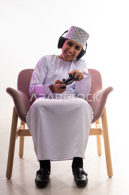 مفهوم الاصغاء والاستمتاع، مواهب غنائية فنية، التفاعل مع الأنغام الموسيقية، الاستماع للأغاني باسخدام الهيدفون، قضاء أوقات ممتعة في ألعاب الفيديو الترفيهيه، بورتريه لولد عربي خليجي عماني يرتدي الدشداشه والكمه يضع سماعات الرأس يمسك بيده عصا التحكم,  خلفيه بيضاء