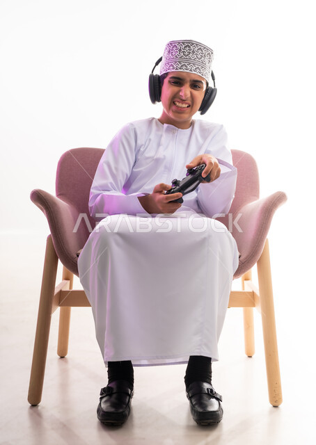 مواهب غنائية فنية، التفاعل مع الأنغام الموسيقية، مفهوم الاصغاء والاستمتاع، الاستماع للأغاني باسخدام الهيدفون، قضاء أوقات ممتعة في ألعاب الفيديو الترفيهيه، بورتريه لولد عربي خليجي عماني يرتدي الدشداشه والكمه يضع سماعات الرأس يمسك بيده عصا التحكم,  خلفيه بيضاء