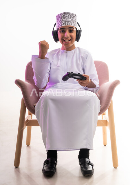 مواهب غنائية فنية، التفاعل مع الأنغام الموسيقية، مفهوم الاصغاء والاستمتاع، الاستماع للأغاني باسخدام الهيدفون، قضاء أوقات ممتعة في ألعاب الفيديو الترفيهيه، بورتريه لولد عربي خليجي عماني يرتدي الدشداشه والكمه يضع سماعات الرأس يمسك بيده عصا التحكم,  خلفيه بيضاء