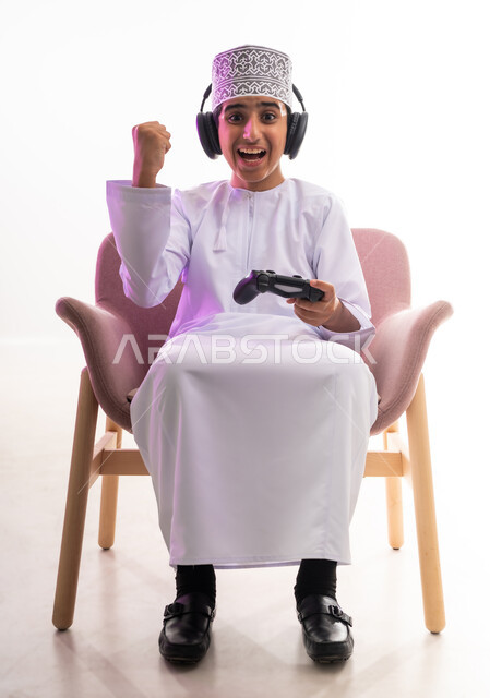 مواهب غنائية فنية، التفاعل مع الأنغام الموسيقية، مفهوم الاصغاء والاستمتاع، الاستماع للأغاني باسخدام الهيدفون، قضاء أوقات ممتعة في ألعاب الفيديو الترفيهيه، بورتريه لولد عربي خليجي عماني يرتدي الدشداشه والكمه يضع سماعات الرأس يمسك بيده عصا التحكم,  خلفيه بيضاء