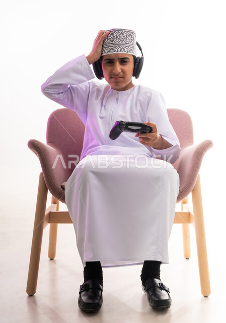 مواهب غنائية فنية، التفاعل مع الأنغام الموسيقية، مفهوم الاصغاء والاستمتاع، الاستماع للأغاني باسخدام الهيدفون، قضاء أوقات ممتعة في ألعاب الفيديو الترفيهيه، بورتريه لولد عربي خليجي عماني يرتدي الدشداشه والكمه يضع سماعات الرأس يمسك بيده عصا التحكم,  خلفيه بيضاء
