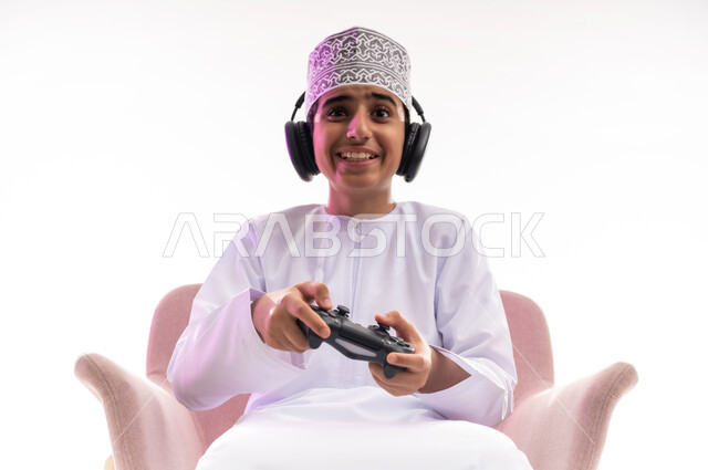 مواهب غنائية فنية، التفاعل مع الأنغام الموسيقية، مفهوم الاصغاء والاستمتاع، الاستماع للأغاني باسخدام الهيدفون، قضاء أوقات ممتعة في ألعاب الفيديو الترفيهيه، بورتريه لولد عربي خليجي عماني يرتدي الدشداشه والكمه يضع سماعات الرأس يمسك بيده عصا التحكم,  خلفيه بيضاء