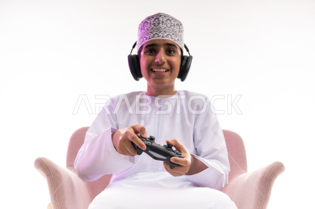 مفهوم الاصغاء والاستمتاع، مواهب غنائية فنية، التفاعل مع الأنغام الموسيقية، الاستماع للأغاني باسخدام الهيدفون، قضاء أوقات ممتعة في ألعاب الفيديو الترفيهيه، بورتريه لولد عربي خليجي عماني يرتدي الدشداشه والكمه يضع سماعات الرأس يمسك بيده عصا التحكم,  خلفيه بيضاء
