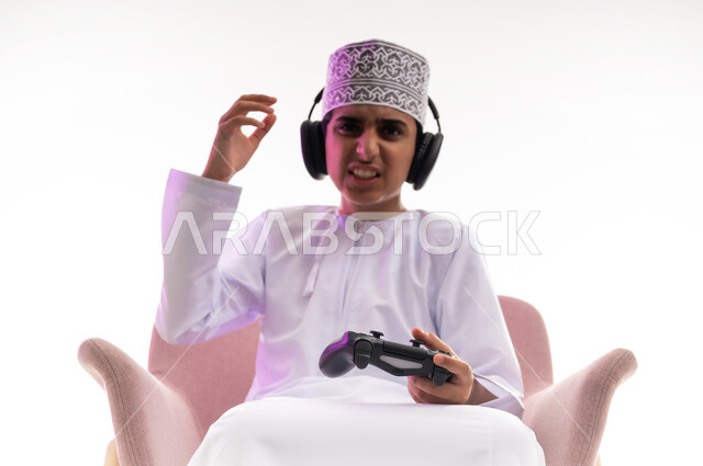 مواهب غنائية فنية، التفاعل مع الأنغام الموسيقية، مفهوم الاصغاء والاستمتاع، الاستماع للأغاني باسخدام الهيدفون، قضاء أوقات ممتعة في ألعاب الفيديو الترفيهيه، بورتريه لولد عربي خليجي عماني يرتدي الدشداشه والكمه يضع سماعات الرأس يمسك بيده عصا التحكم,  خلفيه بيضاء