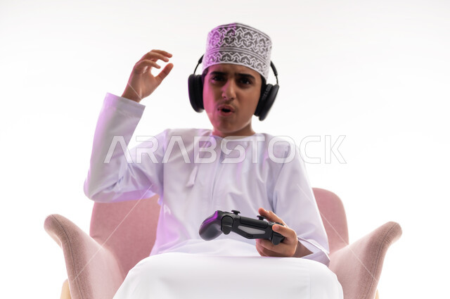 مواهب غنائية فنية، التفاعل مع الأنغام الموسيقية، مفهوم الاصغاء والاستمتاع، الاستماع للأغاني باسخدام الهيدفون، قضاء أوقات ممتعة في ألعاب الفيديو الترفيهيه، بورتريه لولد عربي خليجي عماني يرتدي الدشداشه والكمه يضع سماعات الرأس يمسك بيده عصا التحكم,  خلفيه بيضاء