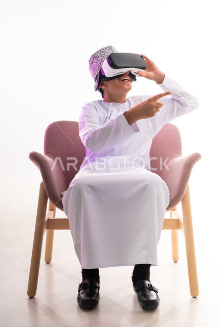 التطور التقني والتكنولوجي، الاندماج ومحاكاة العالم الخيالي والنظر الى الافلام الواقعيه،  بورتريه لولد عربي خليجي عماني يرتدي الدشداشه والكمه ينظر من خلال نظارات ثلاثية الأبعاد (الفي ار) يشير باصبع السبابة الى مكان ما,  الجسم بالطول الكامل، خلفيه بيضاء