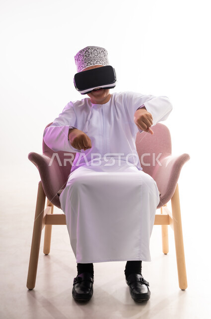 النظر الى الافلام الواقعيه والاندماج ومحاكاة العالم الخيالي ، التطور التقني والتكنولوجي،   بورتريه لولد عربي خليجي عماني يرتدي الدشداشه والكمه ينظر من خلال نظارات ثلاثية الأبعاد (الفي ار) يشير باصبع السبابة الى الاسفل,  الجسم بالطول الكامل، خلفيه بيضاء