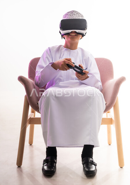 قضاء أوقات ممتعة في ألعاب الفيديو الترفيهيه، الاندماج ومحاكاة العالم الخيالي، استخدام الأجهزة التكنولوجية والتقنيات الحديثة، بورتريه لولد عربي خليجي عماني يرتدي الدشداشه والكمه ينظر من خلال نظارات ثلاثية الأبعاد (الفي ار) يمسك بيده عصا التحكم,  خلفيه بيضاء