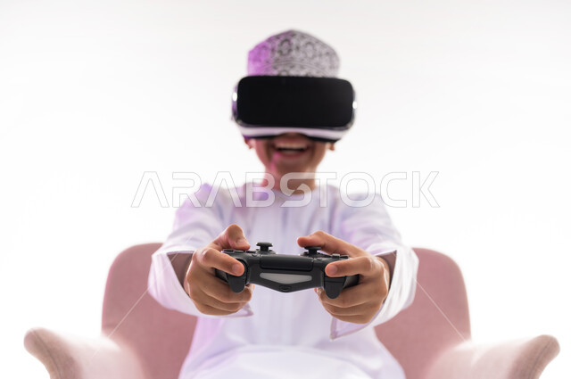 Using technological devices and modern techniques, spending fun times in entertaining video games, merging and simulating the imaginary world, portrait of an Arab Gulf Omani boy wearing a dishdasha and a turban, looking through 3D glasses (VR) holding a joystick in his hand, white background