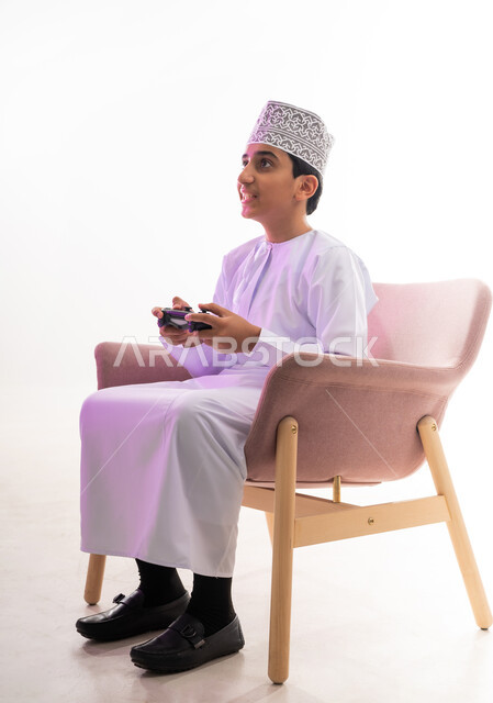 قضاء أوقات ممتعة في ألعاب الفيديو الترفيهيه، بورتريه لولد عربي خليجي عماني يرتدي الدشداشة والكمة يجلس على كرسي مريح يحمل بيده عصا التحكم، إيماءات الفرح والاستمتاع والاندماج، استخدام الأجهزة التكنولوجية والتقنيات الحديثة، خلفية بيضاء