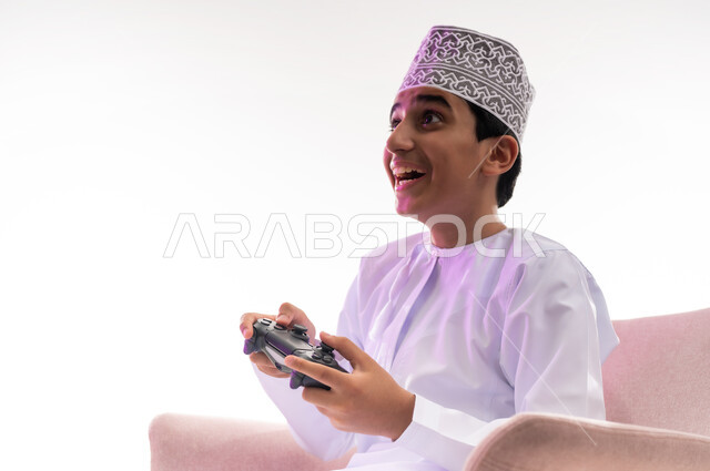 Gestures of joy, enjoyment and integration, use of technological devices and modern techniques, spending enjoyable times playing video games, close-up portrait of an Arab Gulf Omani boy wearing a dishdasha and a kimono sitting on a comfortable chair holding a joystick in his hand, white background
