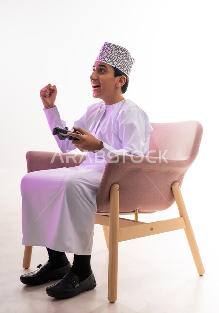 Using technological devices and modern techniques, spending fun times playing entertaining video games, portrait of an Arab Gulf Omani boy wearing a dishdasha and a kimono sitting on a comfortable chair holding a joystick in his hand, gestures of joy, enjoyment and integration, white background