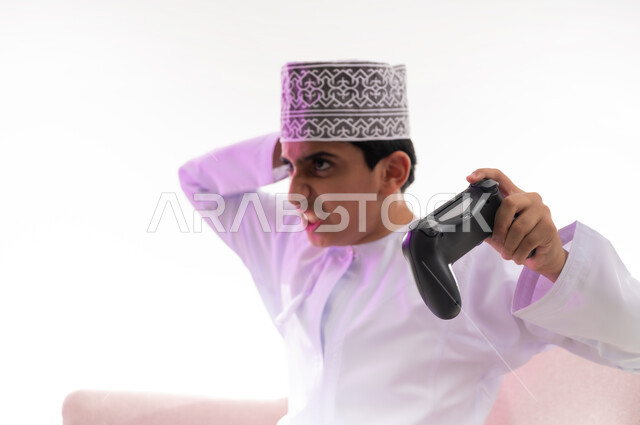 إيماءات الحزن والاندماج،استخدام الأجهزة التكنولوجية والتقنيات الحديثة، قضاء أوقات ممتعة في ألعاب الفيديو الترفيهيه، بورتريه مقرب لولد عربي خليجي عماني يرتدي الدشداشة والكمة يجلس على كرسي مريح يحمل بيده عصا التحكم،  خلفية بيضاء