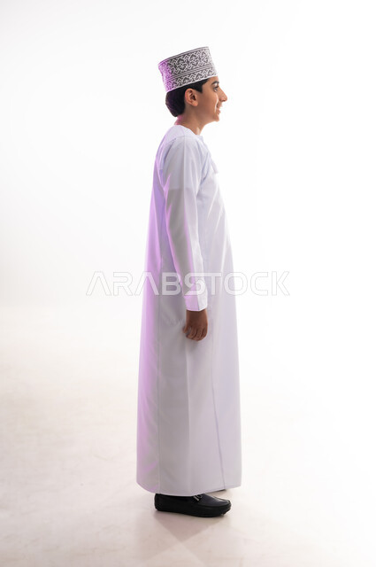 الاهتمام بالأناقة والمظهر الخارجي، مفهوم الرجولة والثقة بالنفس، التقاط الصور الشخصية للوثائق الرسمية، بورتريه من الجانب لولد عربي خليجي عماني مبتسم يرتدي الدشداشة و الكمه بإيماءات السرور والسعادة، الجسم بالطول الكامل، خلفية بيضاء