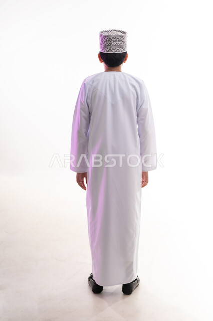 الاهتمام بالأناقة والمظهر الخارجي، بورتريه من الخلف لولد عربي خليجي عماني يرتدي الدشداشة و الكمه ينظر الى شيء ما يقف باستقامة واعتدال، الجسم بالطول الكامل، خلفية بيضاء