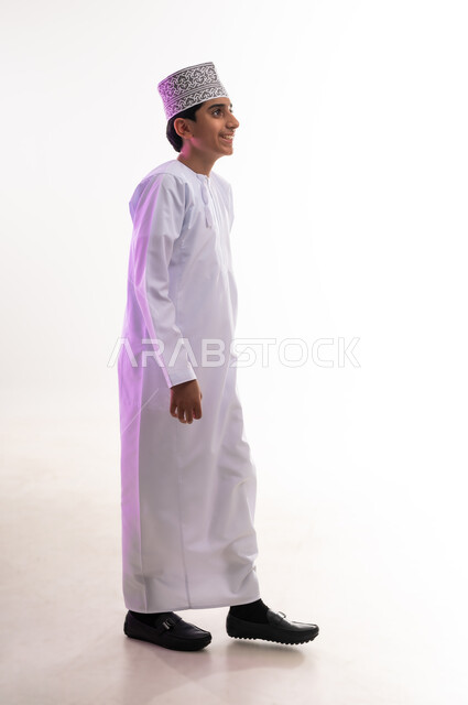 Concern for elegance and appearance, concept of masculinity and self-confidence, taking personal photos for official documents, side portrait of a smiling Omani Gulf Arab boy wearing a dishdasha and a kummah with gestures of pleasure and happiness, full body, white background