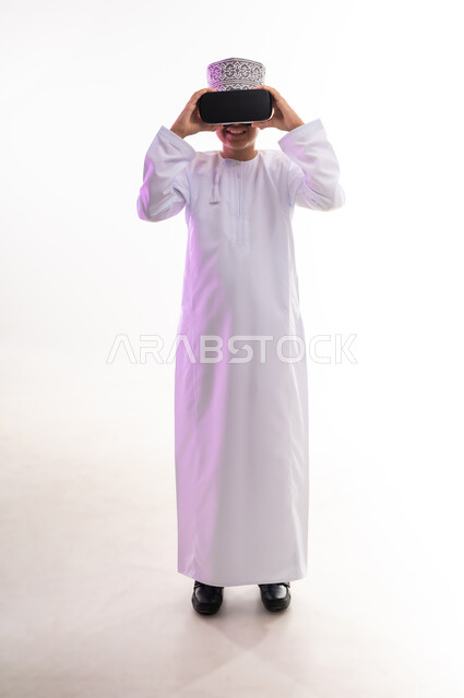 التطور التقني والتكنولوجي, المتعة و التسلية بالمشاهدة عبر نظارة الواقع الافتراضي، بورتريه لولد عربي خليجي عماني يرتدي الدشداشه والكمه ينظر من خلال نظارات ثلاثية الأبعاد (الفي ار), الاندماج ومحاكاة العالم الخيالي والنظر الى الافلام الواقعيه، الجسم بالطول الكامل، خلفيه بيضاء