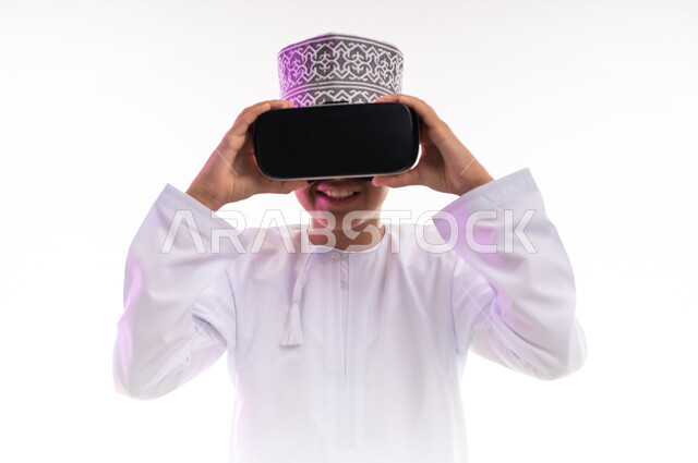 Fun and entertainment by watching through virtual reality glasses, technical and technological development, portrait of an Arab Gulf Omani boy wearing a dishdasha and a cloak looking through 3D glasses (VR), merging and simulating the imaginary world and watching real movies, white background
