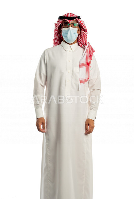 بورتريه لرجل عربي خليجي سعودي ينظر إلى الكاميرا، مرتدي القناع الطبي الوقائي للوقاية من فايروس كورونا كوفيد-19 ، خلفية بيضاء لرجل سعودي بالزي التقليدي السعودي مع شماغ وعقال ، إتباع إجراءات الوقاية من الفايروسات