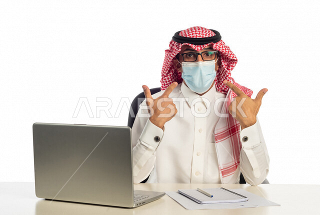 بورتريه لرجل أعمال خليجي سعودي في مقر العمل، يشير بيده إلى الكمامة بإيماءات وجه ويدين مختلفة تدل على إتخاذ إجراءات الوقاية من الأمراض بالعمل، مرتدي القناع الطبي للوقاية من فايروس كورونا كوفيد-19، إستخدام الحاسوب المحمول لإنجاز المهام، بيئة عمل