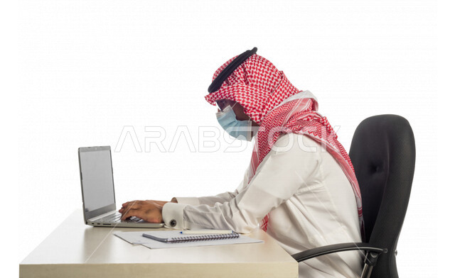 بورتريه لرجل أعمال خليجي سعودي في مقر العمل، مرتدي القناع الطبي للوقاية من فايروس كورونا كوفيد-19، إستخدام الحاسوب المحمول لإنجاز المهام، عقد إجتماع عمل عبر الإنترنت، جلسة عمل،مهن وأعمال ، صفقات إلكترونية، مفهوم إدارة الأعمال، مراجعة البحوث العلمية