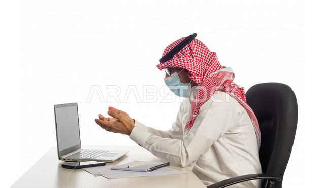 بورتريه لرجل أعمال خليجي سعودي في مقر العمل، مرتدي القناع الطبي الوقائي للوقاية من فايروس كورونا كوفيد-19، إجراء مكالمة فيديو من خلال الحاسوب المحمول لإنجاز المهام، عقد إجتماع عمل عبر الإنترنت، العمل عن بعد، إتباع إجراءات الوقاية، جلسة عمل، أعمال تجارية