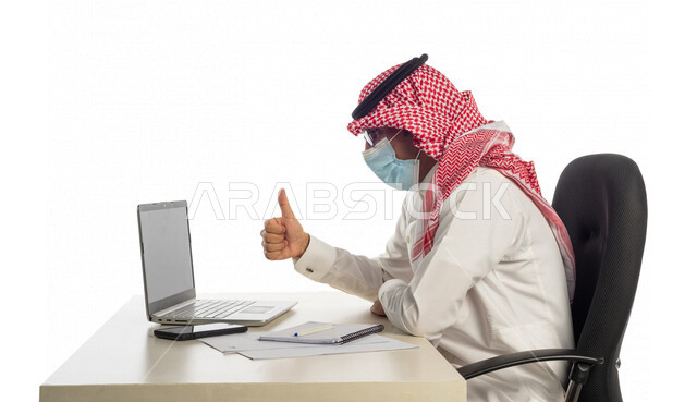 Portrait of a Saudi Gulf businessman at the workplace, wearing a protective medical mask to prevent corona virus Covid-19, making a video call through a laptop to complete tasks, holding a business meeting online, working remotely, following prevention me