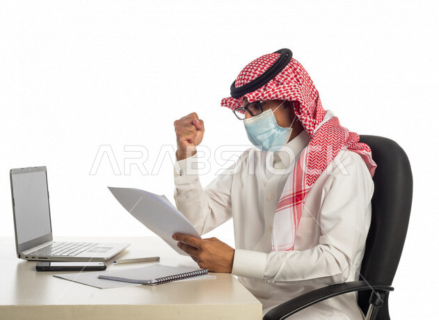 بورتريه لرجل أعمال خليجي سعودي في مقر العمل، مرتدي القناع الطبي الوقائي للوقاية من فايروس كورونا كوفيد-19، إجراء مكالمة فيديو من خلال الحاسوب المحمول لإنجاز المهام، عقد إجتماع عمل عبر الإنترنت، العمل عن بعد، إتباع إجراءات الوقاية، جلسة عمل، أعمال تجارية