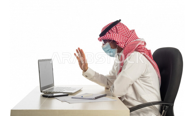 Portrait of a Saudi Gulf businessman at the workplace, wearing a protective medical mask to prevent corona virus Covid-19, making a video call through a laptop to complete tasks, holding a business meeting online, working remotely, following prevention me