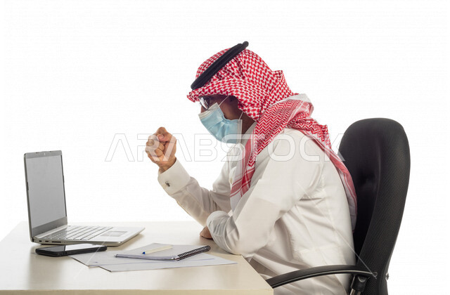 بورتريه لرجل أعمال خليجي سعودي في مقر العمل، مرتدي القناع الطبي الوقائي للوقاية من فايروس كورونا كوفيد-19، إجراء مكالمة فيديو من خلال الحاسوب المحمول لإنجاز المهام، عقد إجتماع عمل عبر الإنترنت، العمل عن بعد، إتباع إجراءات الوقاية، جلسة عمل، أعمال تجارية