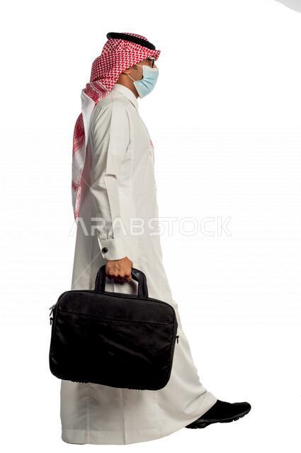 Portrait of a Saudi Gulf businessman carrying a bag to go to work with different facial and hand gestures, wearing a protective medical mask to prevent corona virus COVID-19, side view of a Saudi man in traditional Saudi dress, following virus prevention