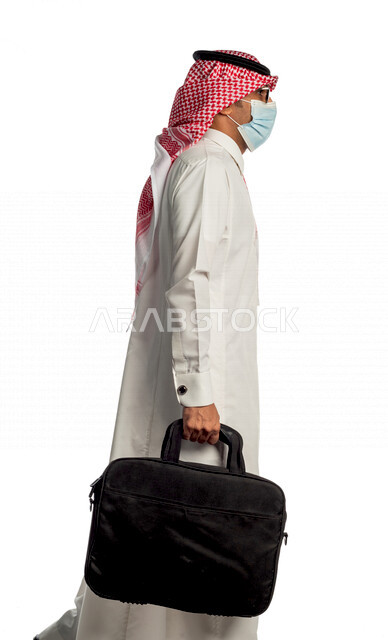 Portrait of a Saudi Gulf businessman carrying a bag to go to work with different facial and hand gestures, wearing a protective medical mask to prevent corona virus COVID-19, side view of a Saudi man in traditional Saudi dress, following virus prevention