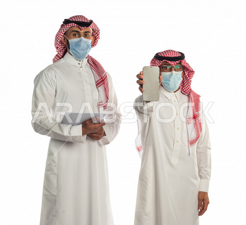 بورتريه لموظفان خليجيان سعوديان، رجل أعمال يحمل أوراق خطة العمل، والأخر يحمل الهاتف المحمول، عرض خدمات وتطبيقات خاصة بالعمل، إرتداء القناع الطبي للوقاية من فايروس كورونا كوفيد-19،إعلان وتسويق، متابعة إستراتيجية العمل، إستخدام أجهزة تقنية حديثة