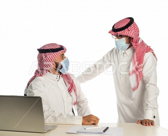 بورتريه لموظفان خليجيان سعوديان في مقر العمل، إرتداء القناع الطبي الوقائي للوقاية من فايروس كورونا كوفيد-19، إستخدام الحاسوب المحمول لإنجاز المهام، العمل عن بعد، زملاء عمل، شركة سعودية،أعمال تجارية، إتباع إجراءات الوقاية، مفهوم إدارة الأعمال، مناقشة العمل