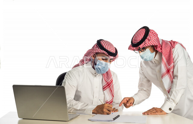بورتريه لموظفان خليجيان سعوديان في مقر العمل، إرتداء القناع الطبي الوقائي للوقاية من فايروس كورونا كوفيد-19، إستخدام الحاسوب المحمول لإنجاز المهام، العمل عن بعد، زملاء عمل، شركة سعودية،أعمال تجارية، مفهوم إدارة الأعمال، مناقشة العمل، مراجعة البحوث العلمية