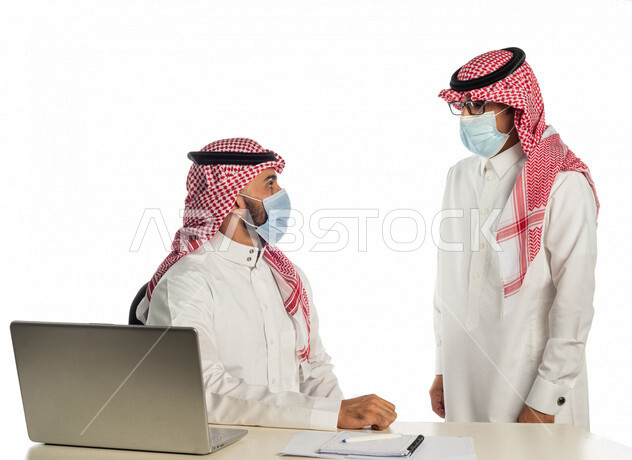 بورتريه لموظفان خليجيان سعوديان في مقر العمل، إرتداء القناع الطبي الوقائي للوقاية من فايروس كورونا كوفيد-19، إستخدام الحاسوب المحمول لإنجاز المهام، العمل عن بعد، زملاء عمل، شركة سعودية،أعمال تجارية، مفهوم إدارة الأعمال، مناقشة العمل، مراجعة البحوث العلمية