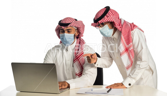بورتريه لموظفان خليجيان سعوديان في مقر العمل، إرتداء القناع الطبي الوقائي للوقاية من فايروس كورونا كوفيد-19، إستخدام الحاسوب المحمول لإنجاز المهام، العمل عن بعد، زملاء عمل، شركة سعودية،أعمال تجارية، مفهوم إدارة الأعمال، مناقشة العمل، مراجعة البحوث العلمية